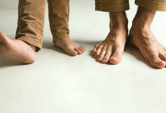 Adult and child standing barefoot side by side in matching khaki pants, representing the cold-sensitive feet that Raynaud's syndrome affects.