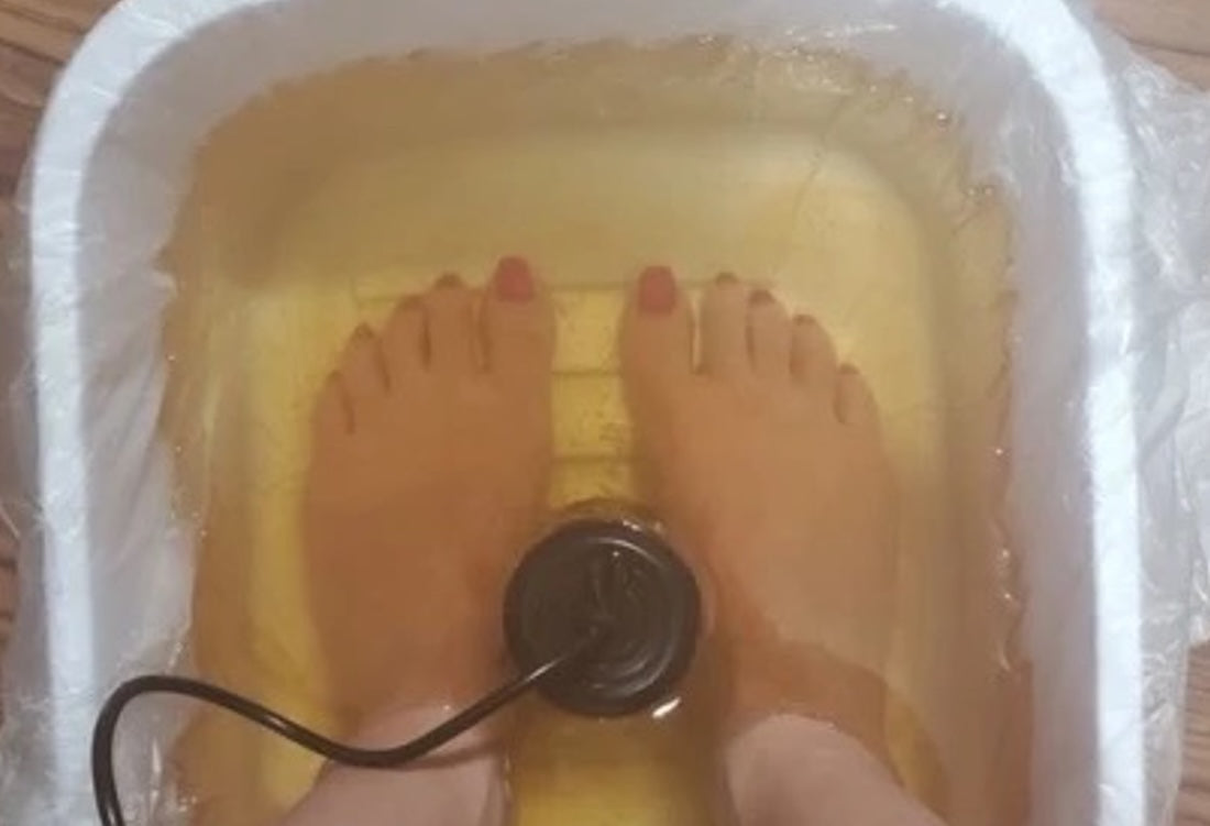 Person soaking both feet in a disposable plastic-lined foot bath basin during an ionic foot spa session, with the ionizer array positioned between the feet. 