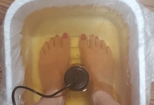 Person soaking both feet in a disposable plastic-lined foot bath basin during an ionic foot spa session, with the ionizer array positioned between the feet. 